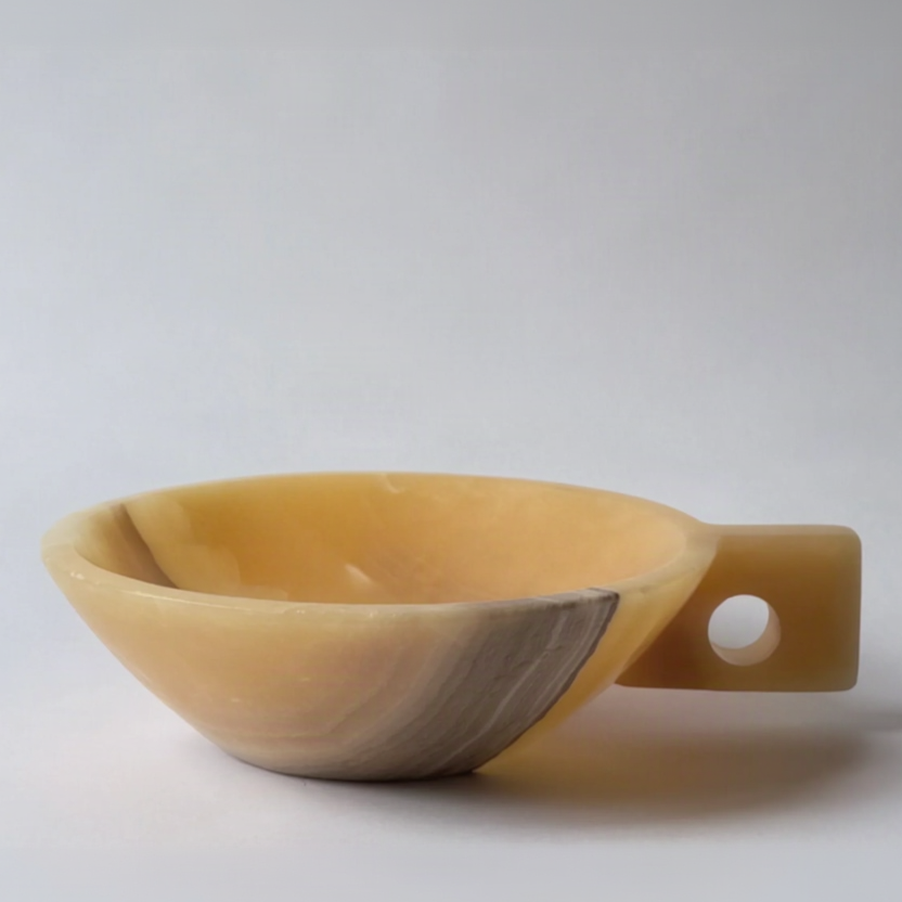 A small handcrafted stone bowl with a handle and circular cutout, made from marbled beige and gray agate or onyx, photographed on a light gray background.